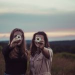 Two ladies holding daisy's in front of their face.